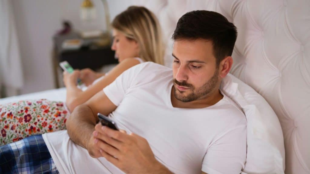 A man and woman are lying in bed, focused on their separate mobile phones.