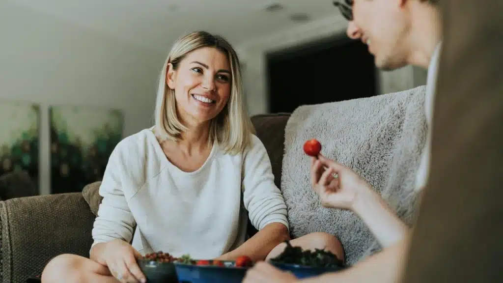 A woman smiling while sitting on a couch and sharing food with a man.