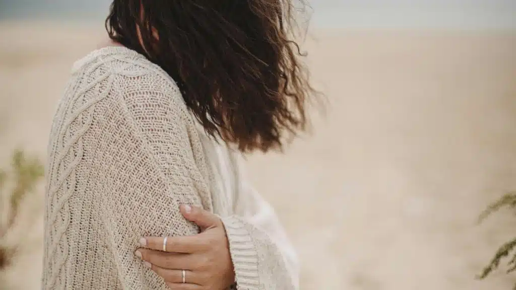A woman in a knit sweater stands on a beach with windblown hair.