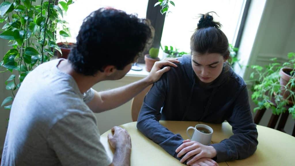 A man comforts a seated woman with a hand on her shoulder at a table with a mug.