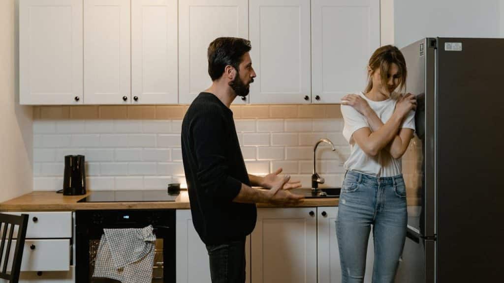 Man with a beard gesturing while talking to a woman with crossed arms in a modern white kitchen.