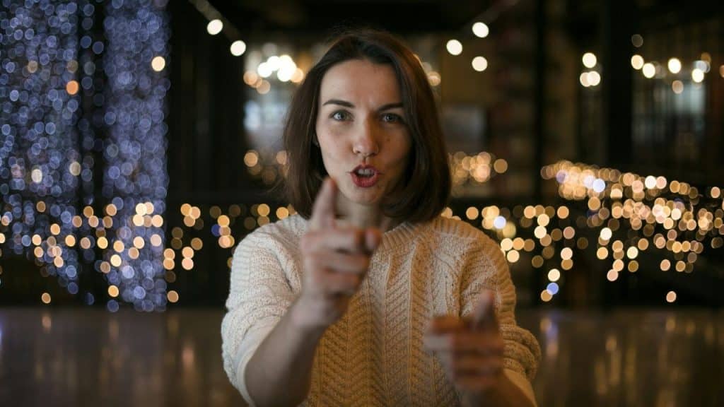 Woman with bobbed hair pointing two fingers forward in front of blurred lights.