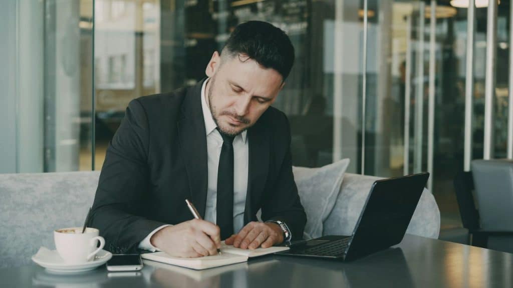 Man in a black suit and tie writing in a notebook at a table with a laptop and coffee.