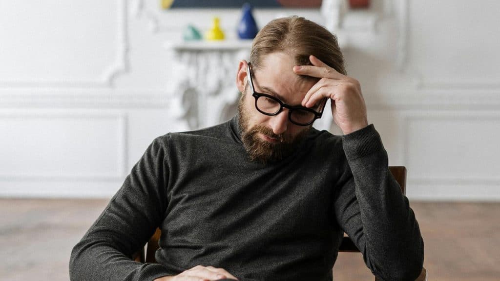 A man wearing glasses sitting with his hand on his forehead, looking thoughtful.