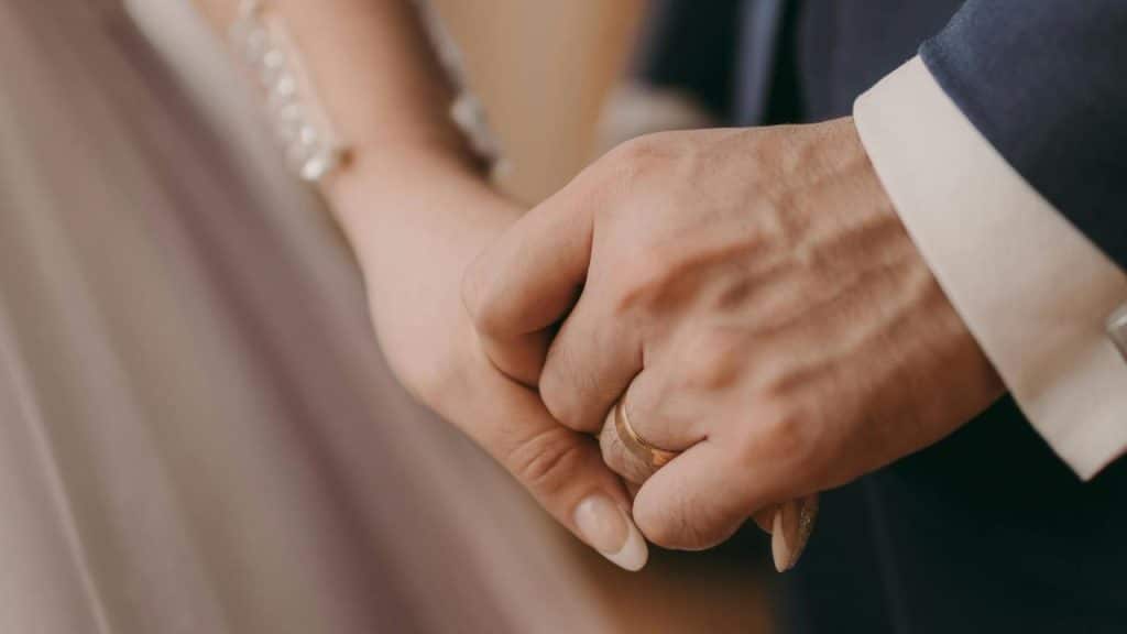 A close-up of a bride and groom holding hands, showing their wedding rings.