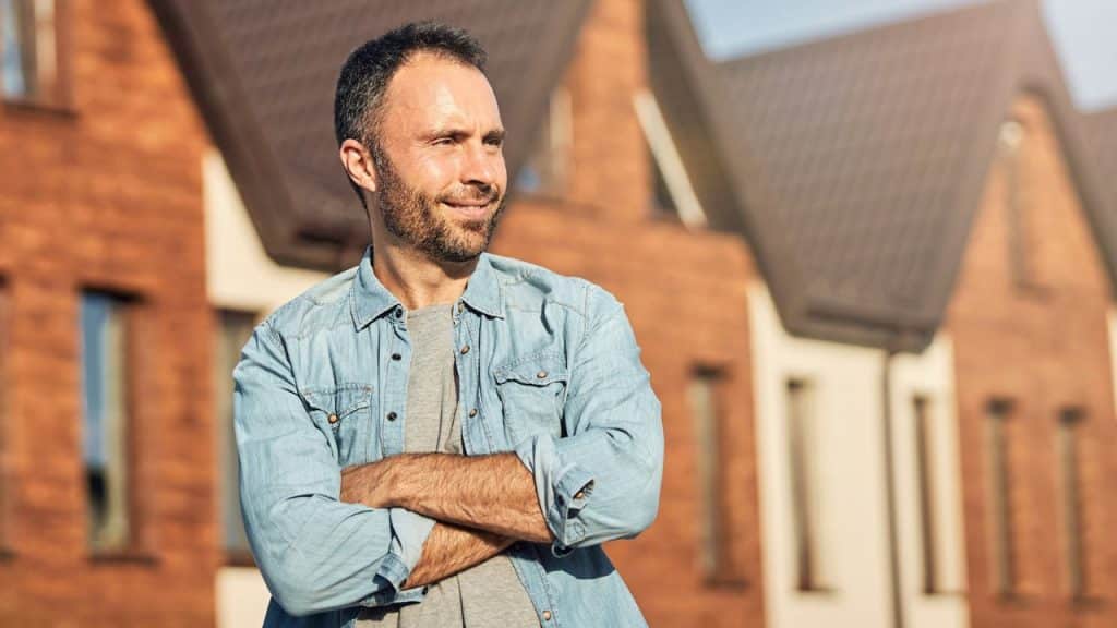Smiling man with a beard and denim shirt, arms crossed, standing outside houses.