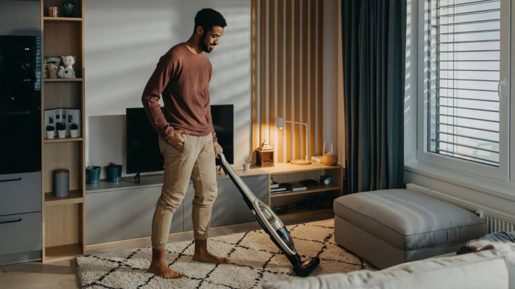 Smiling man vacuuming a patterned rug in a modern, well-lit living room.