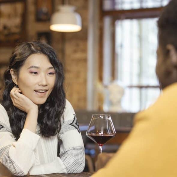 Woman with long, dark hair talking to a man over a glass of red wine in a dimly lit restaurant