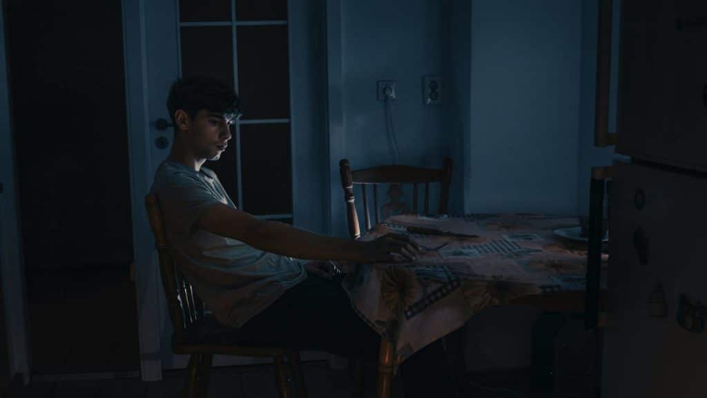 Young man sitting at a kitchen table in a dim, dark, blue-lit room.