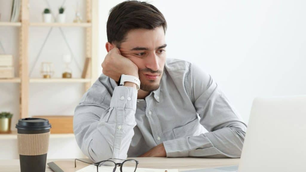 A bored-looking man sits at a desk, resting his head on his hand in front of a laptop.