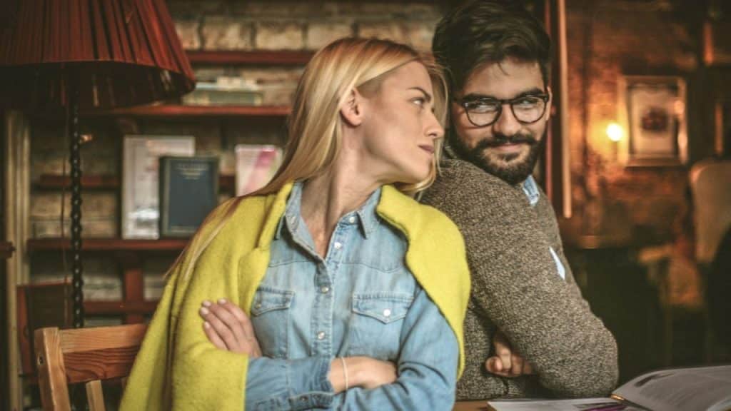 Blonde woman and bearded man with glasses looking at each other indoors.