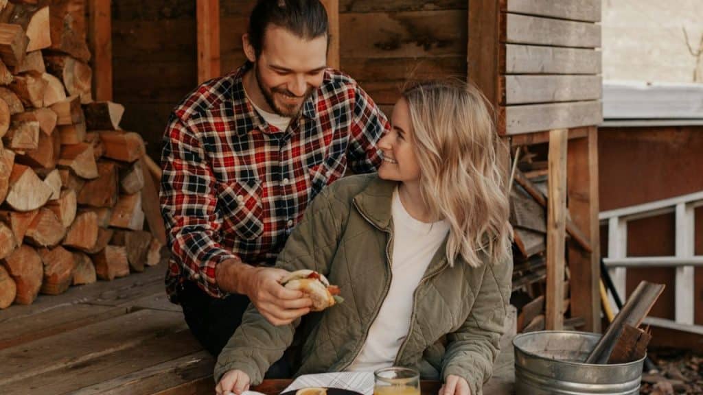 A smiling man in a plaid shirt offers a sandwich to a smiling woman sitting outside a cabin.