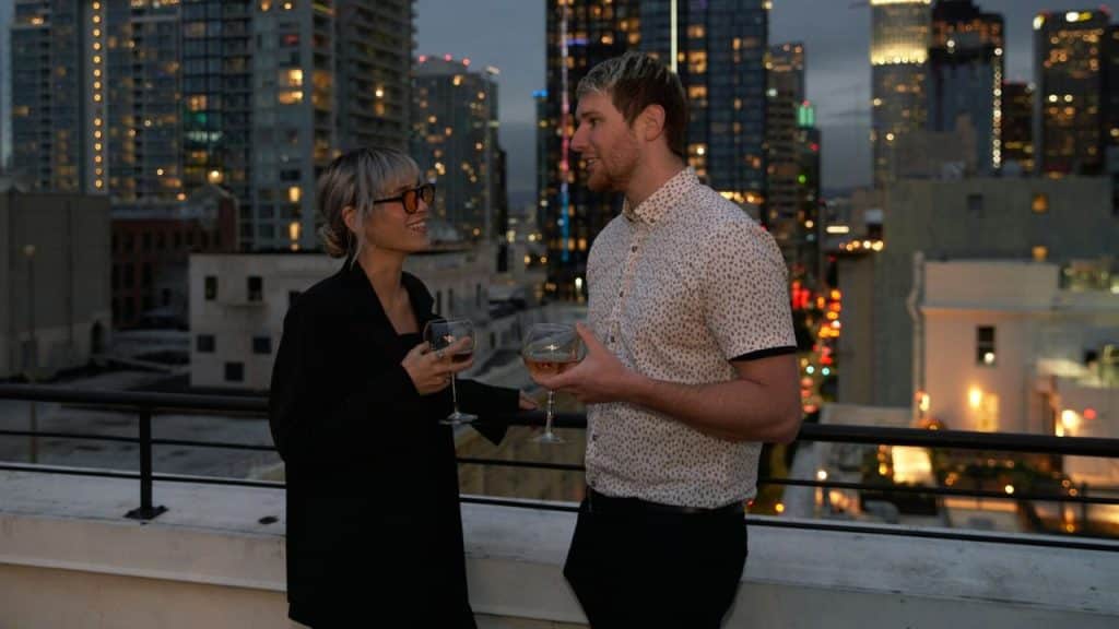 A man and a woman are standing on a rooftop at night, holding wine glasses and talking with a city skyline in the background.