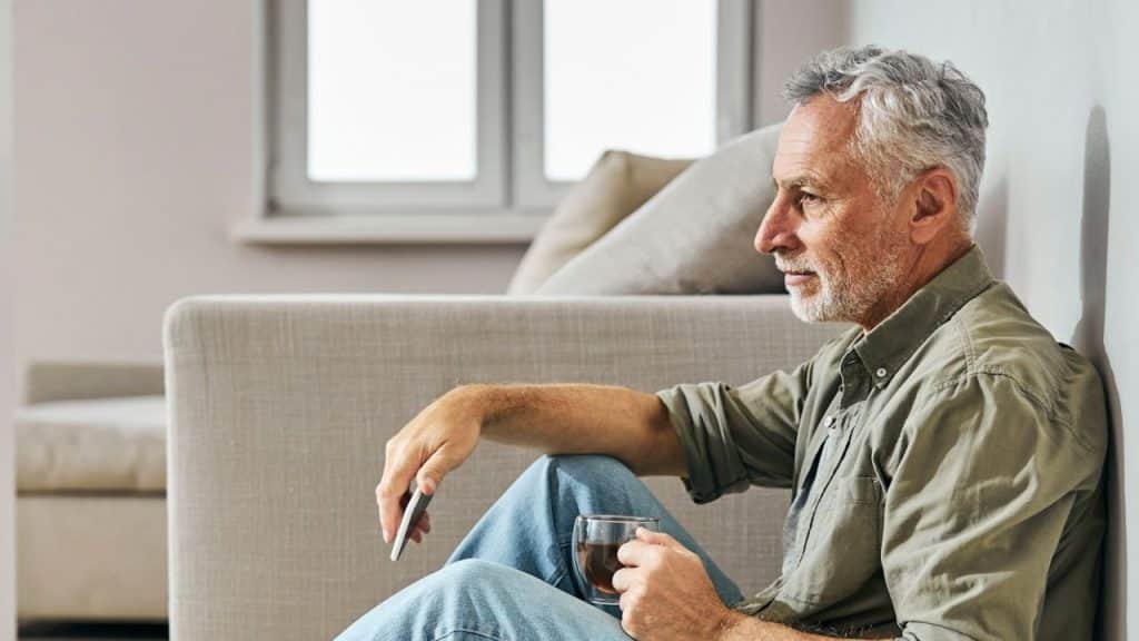 An older man with gray hair and a beard is sitting on the floor, leaning against a wall, holding a phone and a glass.