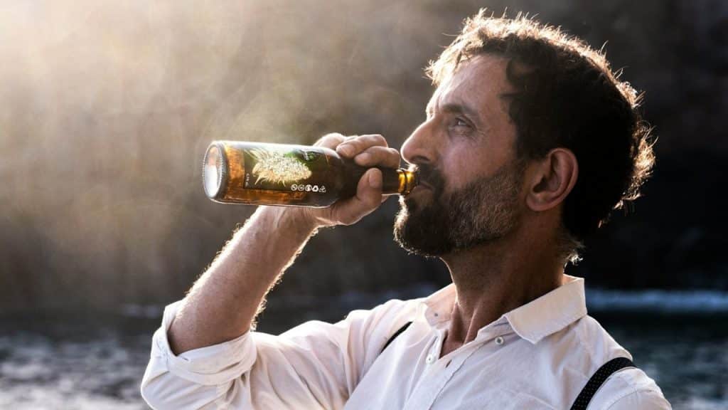 A middle-aged man with a beard is drinking from a glass bottle in the sunlight.