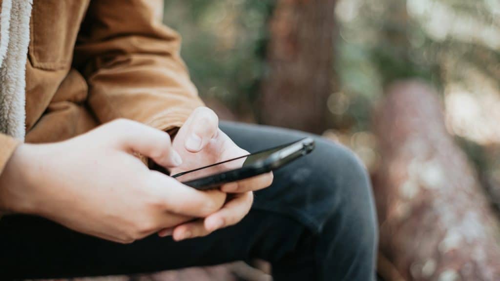 Close-up of a person wearing a brown corduroy jacket with a sherpa collar and black pants, sitting outdoors and texting on a smartphone.