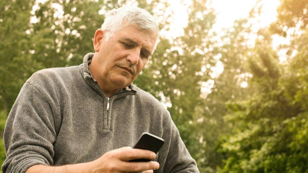 Older man in a gray sweater looking down at his smartphone outdoors with trees.