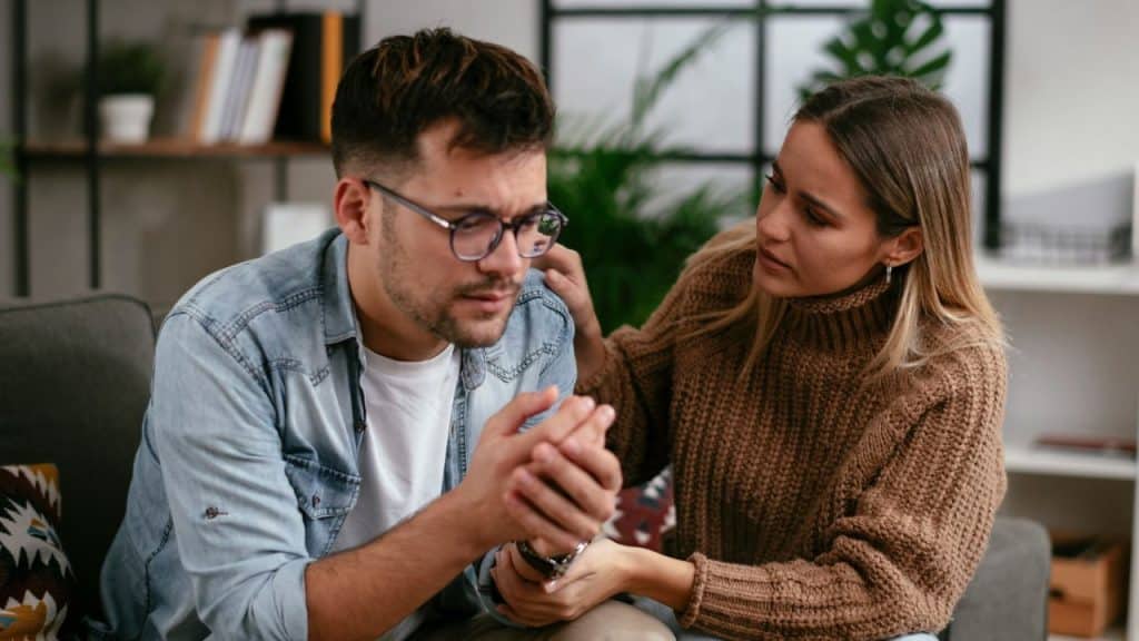 Woman gently touching and consoling a man wearing glasses who looks distressed.