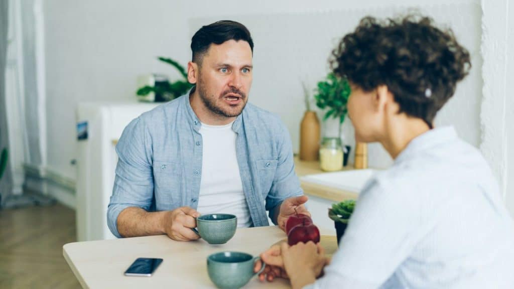 Man looking shocked and speaking intensely to a woman across a table with coffee cups.