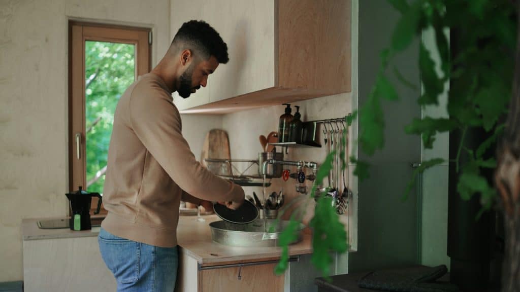 A man with a beard, wearing a beige sweatshirt and blue jeans, is washing dishes in a sink in a bright, modern wooden kitchen.