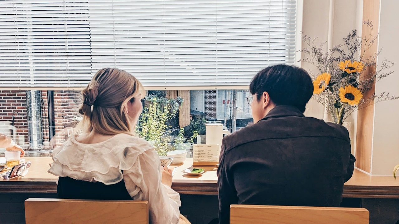 A woman and a man sit facing a window with closed blinds at a table, with a small vase of sunflowers to the right.