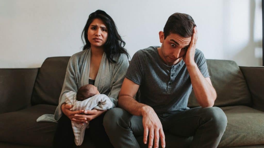 A stressed couple sits on a couch, the woman holding a newborn baby, both looking upset.