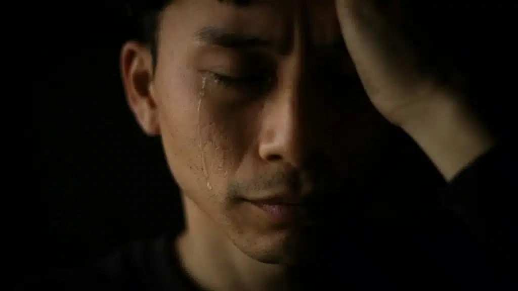 Close-up of a man in darkness with his eyes closed and a tear running down his cheek.