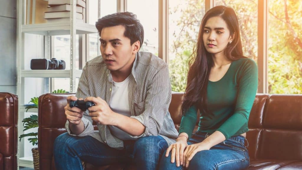A man plays a video game on a couch next to a woman who looks bored or sad.