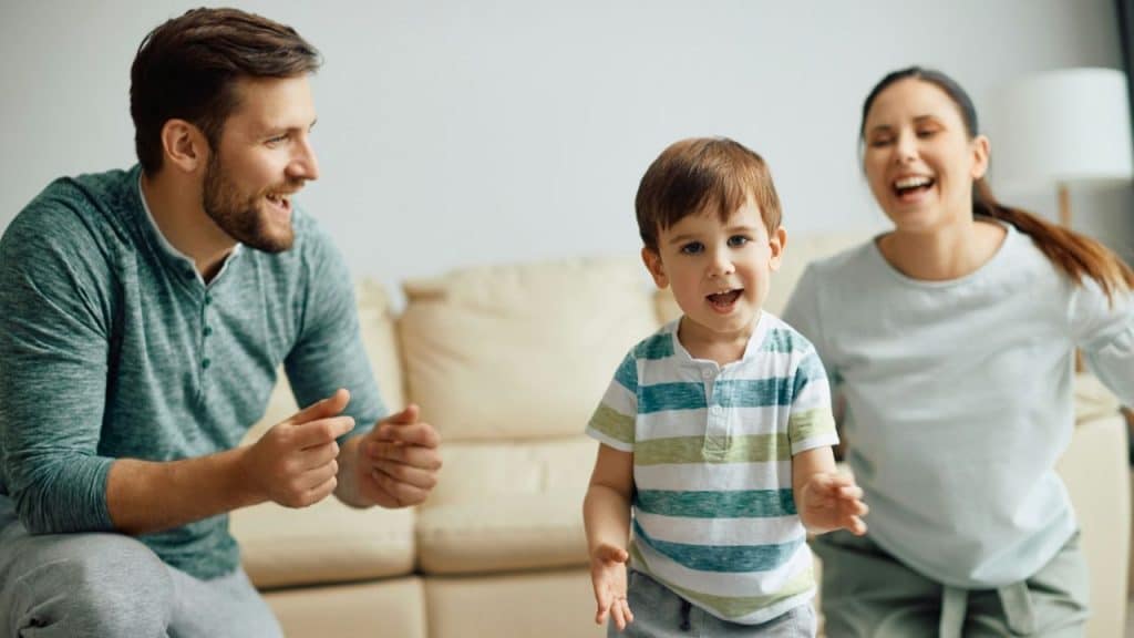 A toddler smiles at the camera while his smiling parents play with him in a living room.