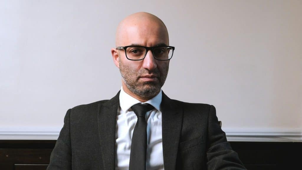 Bald man with a beard, glasses, and a suit staring intensely at the camera.
