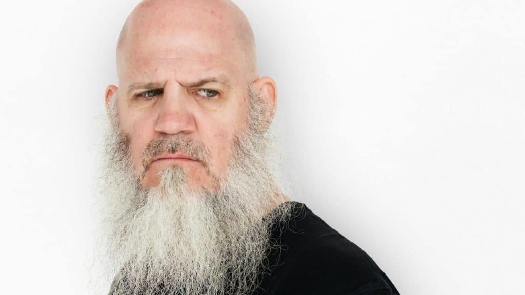 Bald man with a long white beard looking serious against a white background.