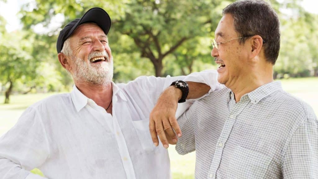 Two older men smiling and laughing together in a sunny park setting.
