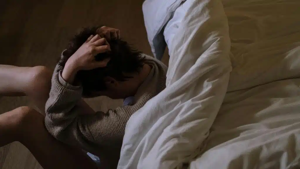 A person sitting on the floor beside a bed with their head in their hands.