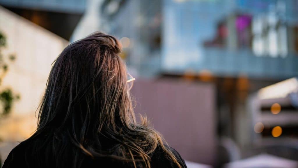 A person with long hair viewed from behind, looking toward a blurred modern building.