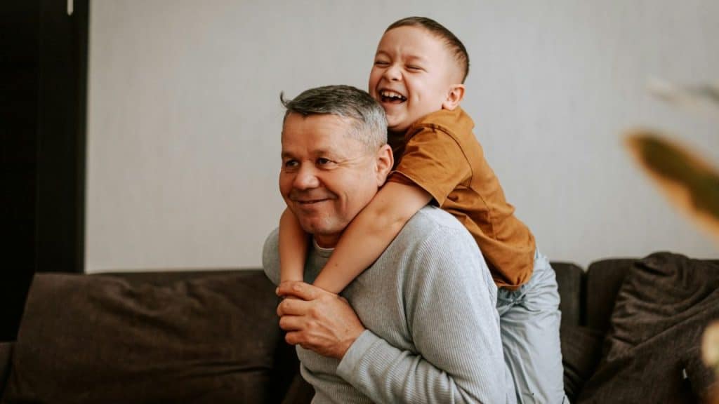 Happy child laughing while getting a piggyback ride from an older man on a sofa.