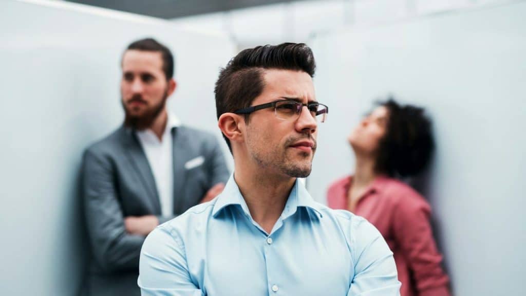 Man in a blue shirt and glasses looking away, with two blurred people behind him.