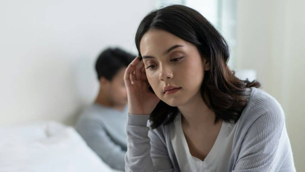 Woman sitting in foreground with hand near her head, and a person in the background out of focus.