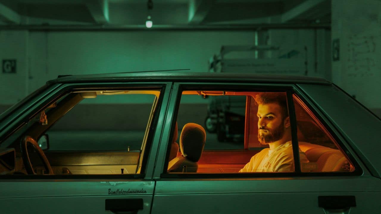 Bearded man sitting in a car at night with the interior dimly lit by orange light.