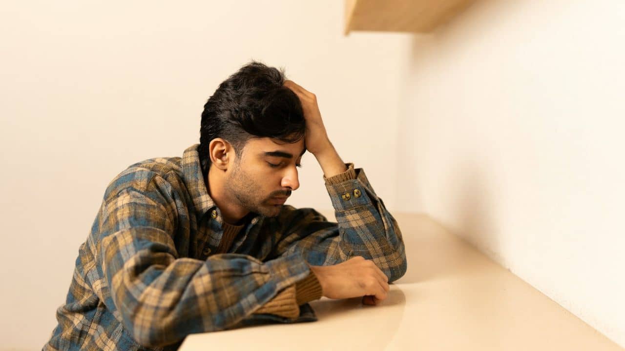 Man in a plaid shirt resting his head on his hand at a table, looking stressed.