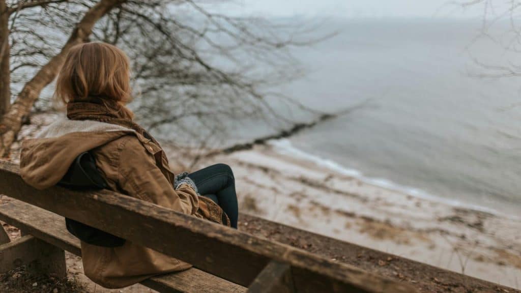 A person wearing a brown jacket sits on a bench overlooking a calm beach and sea.