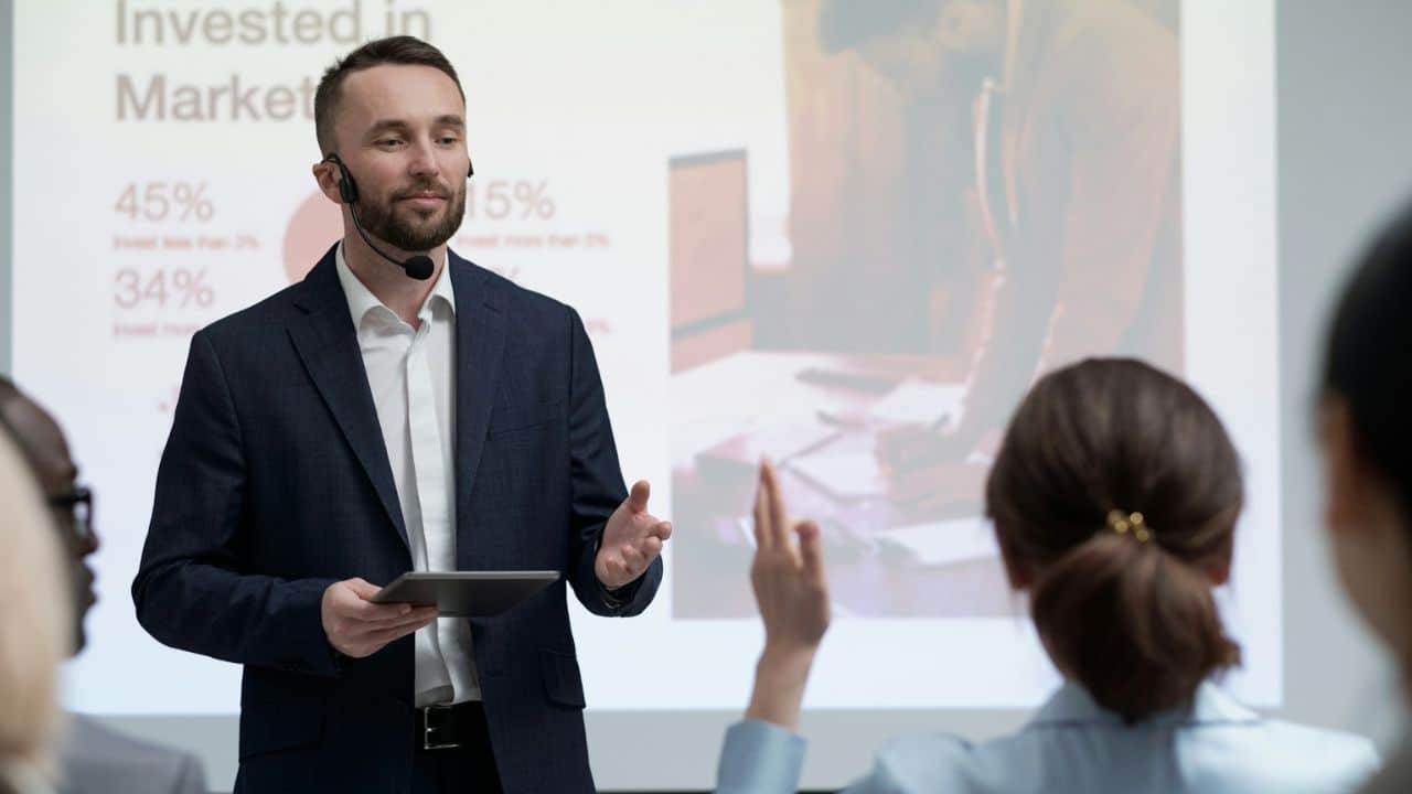 Man with a headset and tablet presenting to an audience with a slide projection behind him.
