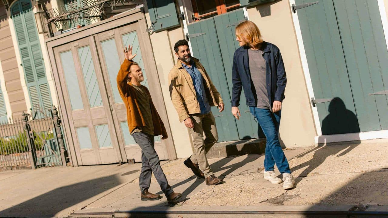 Three men talking and walking down a sunny street with blue and gray buildings.