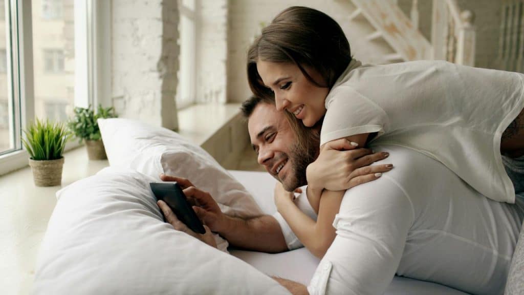A smiling couple lies on a bed watching something on a phone together.