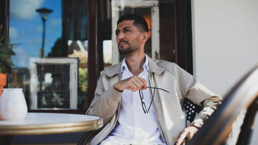Man in a light jacket and white shirt sitting at an outdoor table, holding glasses.