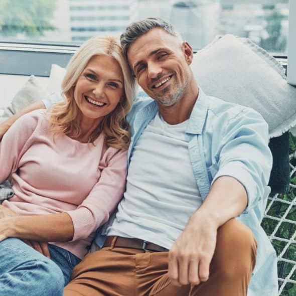 Smiling middle-aged couple relaxing together on a woven hammock indoors by a window.