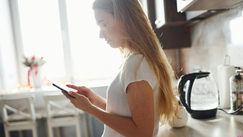 A woman standing in a bright kitchen while using her phone.