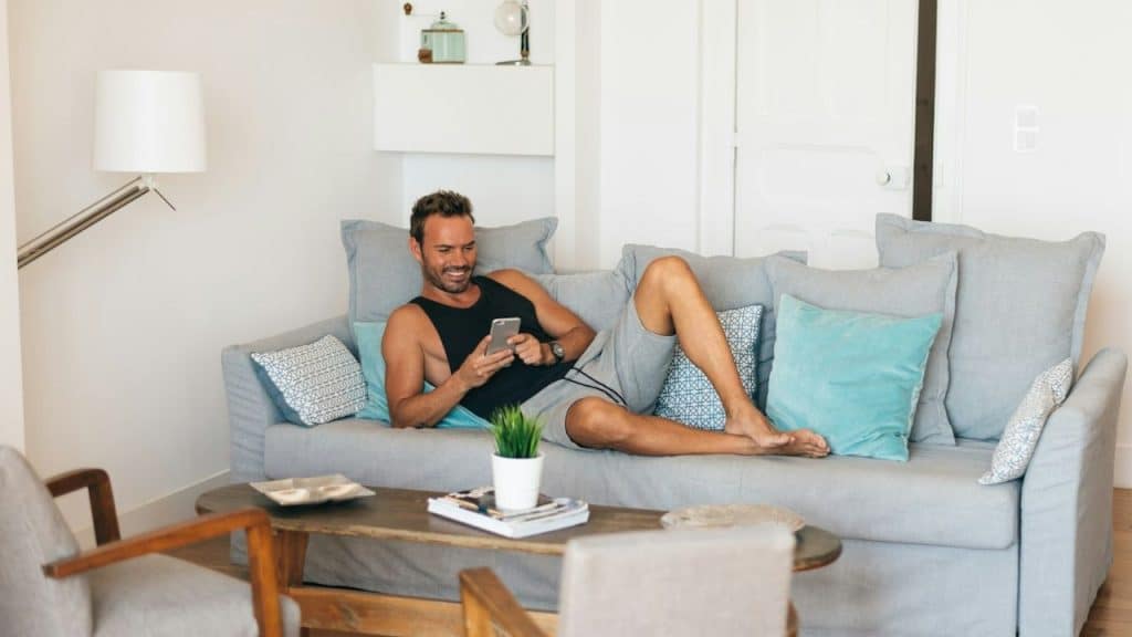 Man lounging on a gray couch, looking at his phone, in a bright living room.