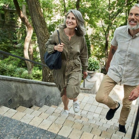 A smiling older couple walks up outdoor stone steps surrounded by greenery.