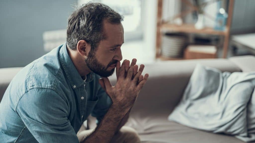 A bearded man in a denim shirt sits on a couch, looking worried with clasped hands.