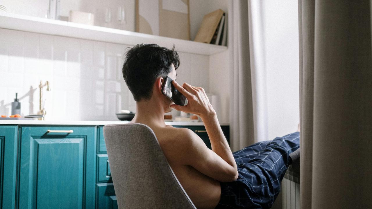 A man without a shirt making a phone call, with his feet raised.